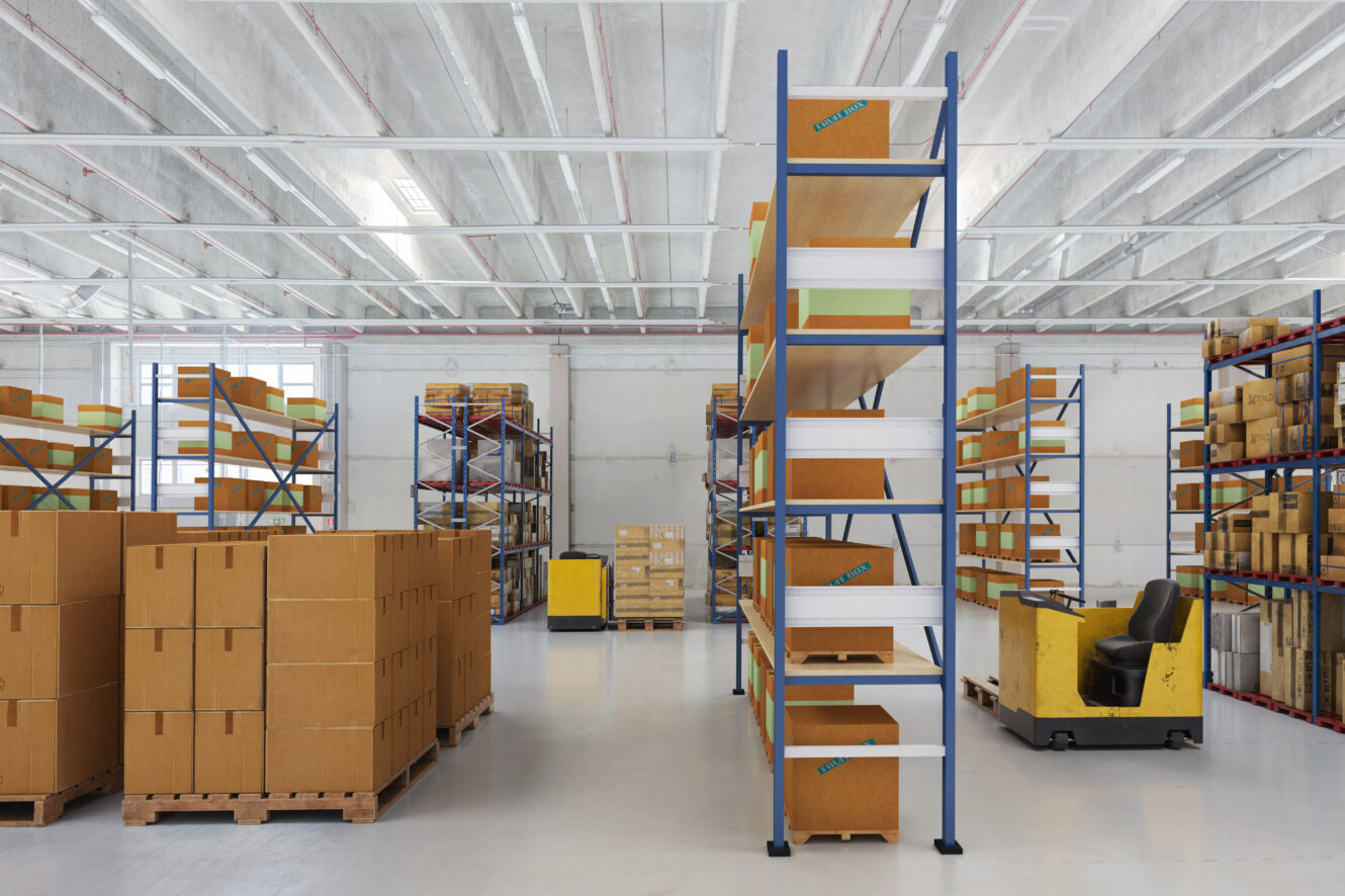 Varaston sisätiloissa on korkeita hyllyjä täynnä pahvilaatikoita, kuormalavoja lattialla ja keltainen trukki pysäköitynä oikealla puolella.