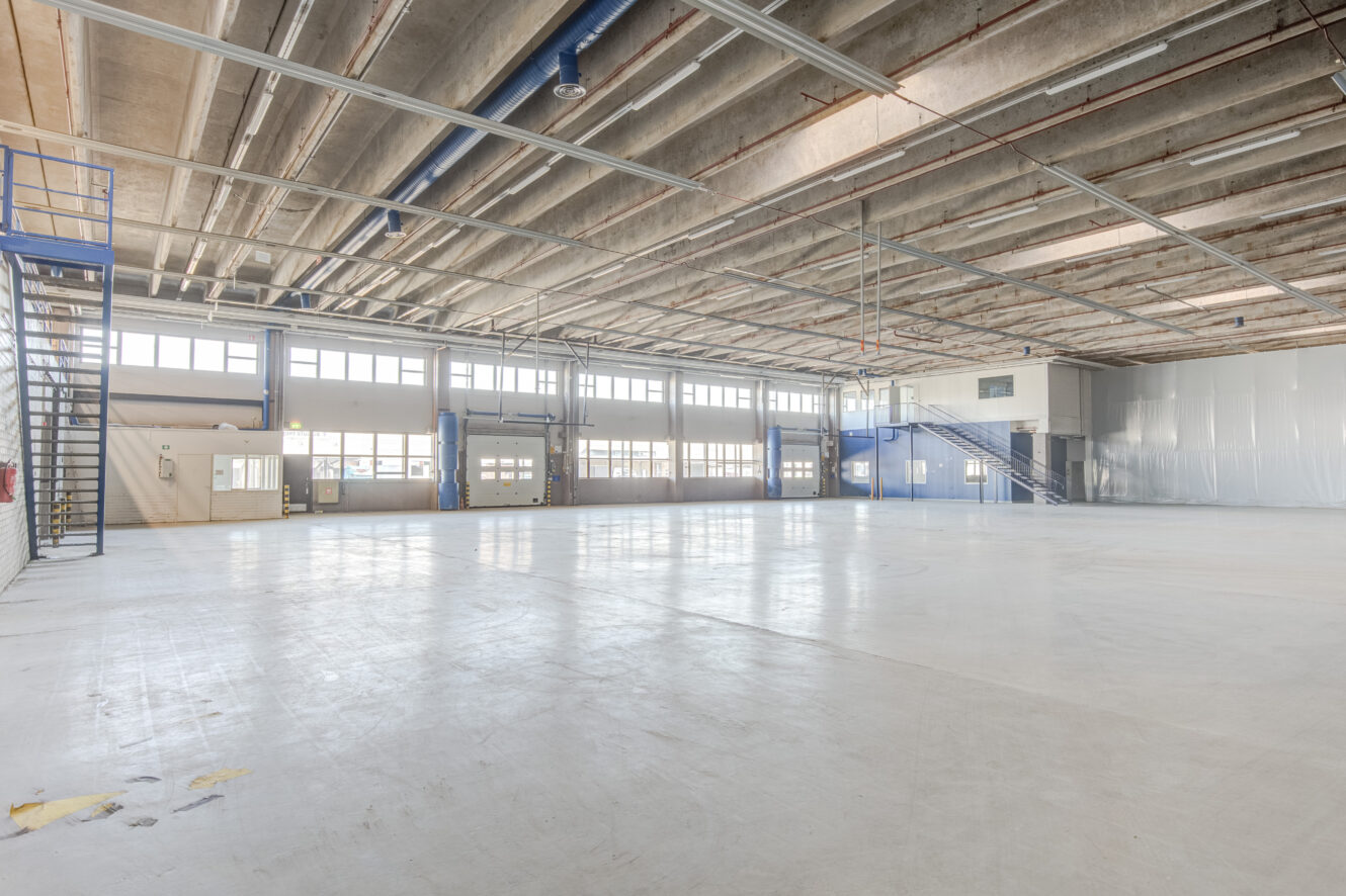 Tilava, tyhjä varaston sisätila, jossa on suuret ikkunat, betonilattia, paljaat kattoputket ja portaat, jotka johtavat ylemmille tasanteille molemmin puolin.