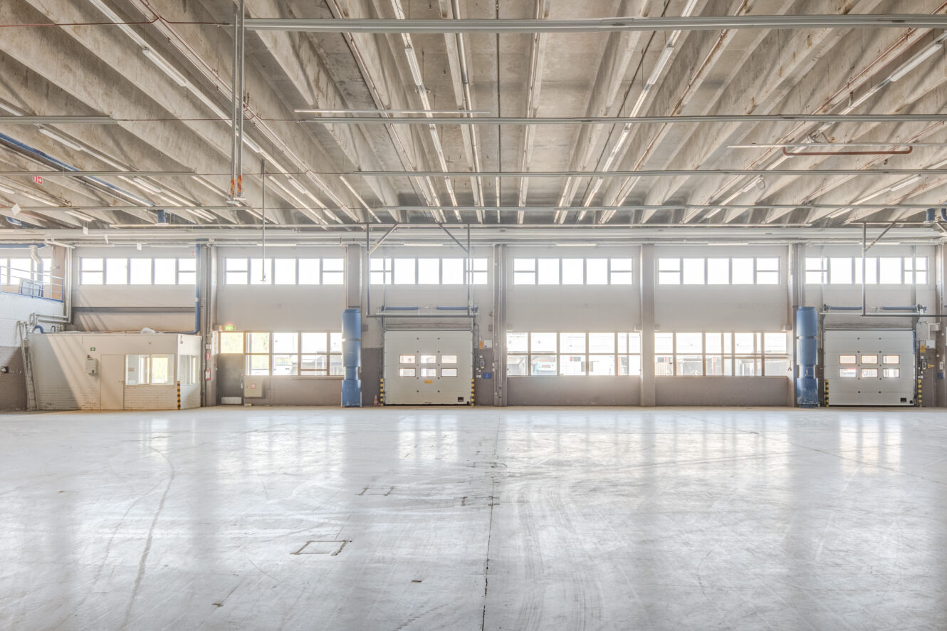 Tyhjä teollisuusvarasto, jossa on betonilattia, korkea katto, suuret ikkunat, lastauslaiturit ja pieni toimistorakennus vasemmalla puolella.