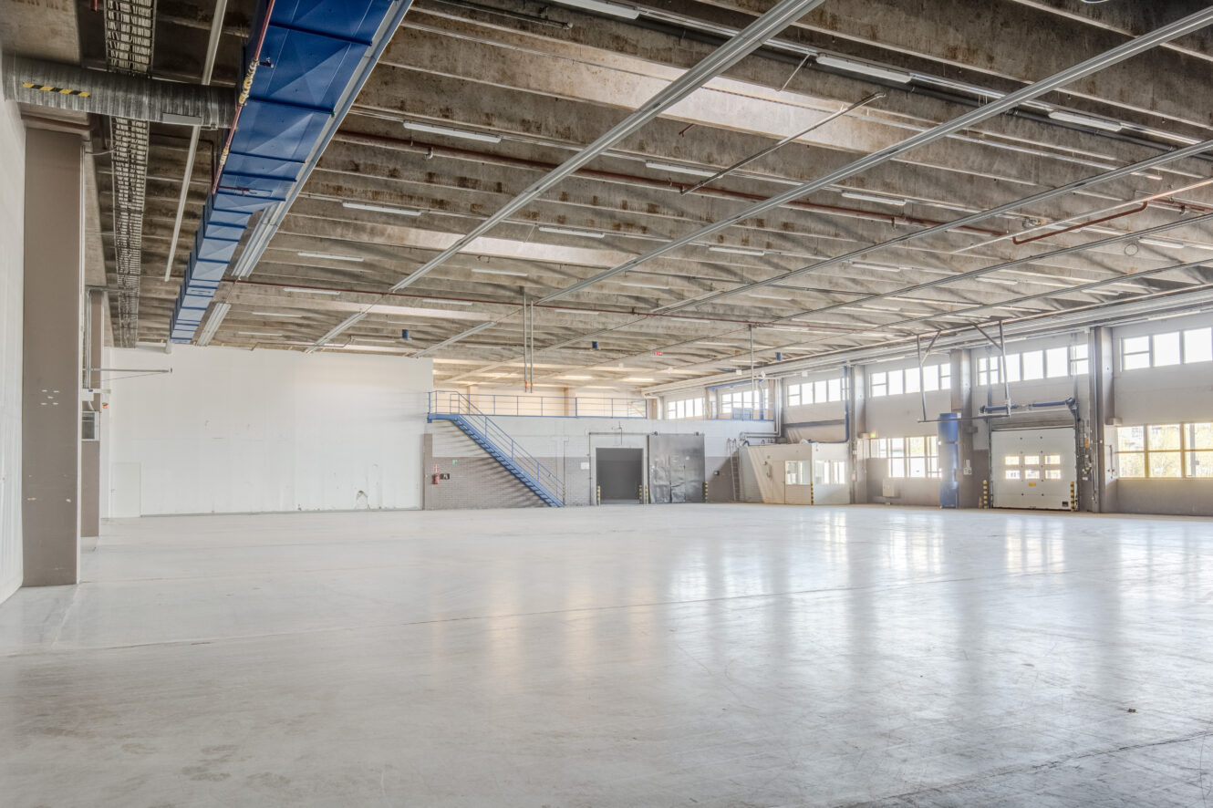 Tilava, tyhjä teollisuusvarasto, jossa on korkeat katot, palkit, suuret ikkunat ja kaksi lastauslaiturin ovea; portaikko johtaa vasemmalla olevaan välipohjatasoon.