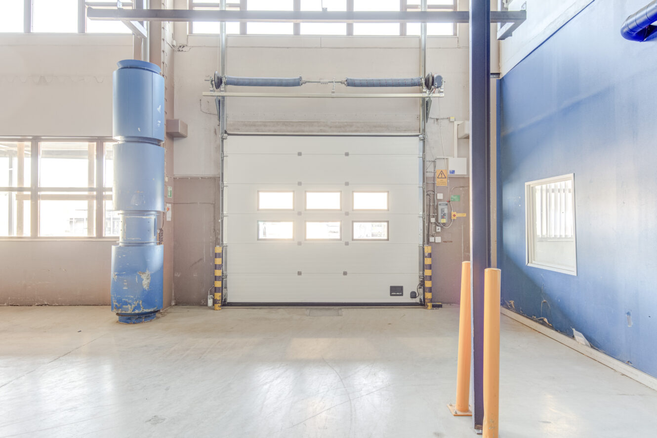 Teollinen sisätila, jossa on suljettu valkoinen autotallin yläovi, vasemmalla siniset putket, turvapollarit ja oikealla sininen seinä, jossa on ikkuna.