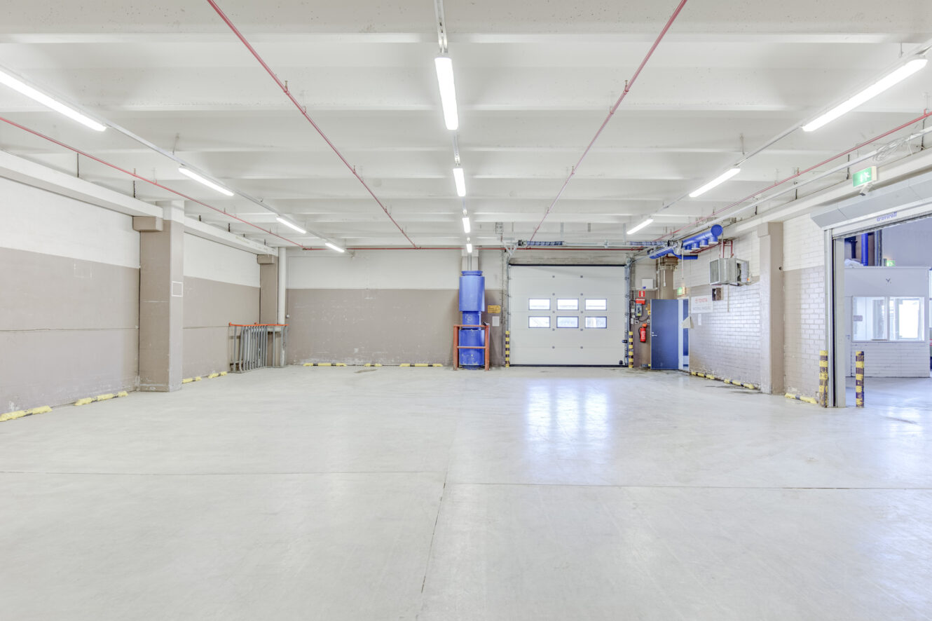 Tyhjä teollisuusvarasto, jossa on valkoiset seinät, kirkas ylävalaistus, suuri autotallin ovi ja turvamerkinnät seinien alaosassa.