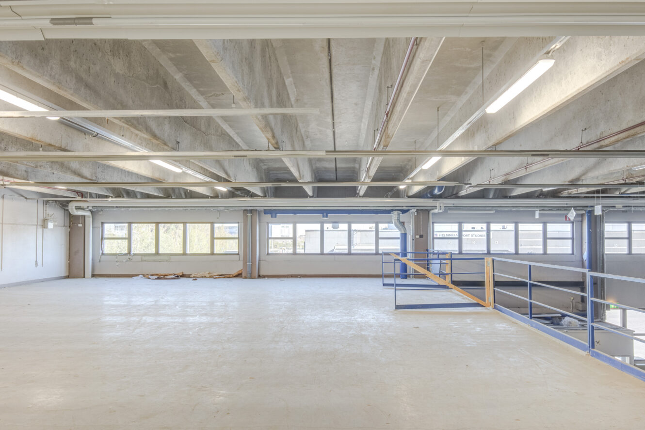 Tyhjä teollinen parvitila, jossa on betonilattiat, paljaat kattopalkit ja suuret ikkunat, jotka antavat luonnonvaloa; sininen kaide kulkee korotetun alustan reunalla.