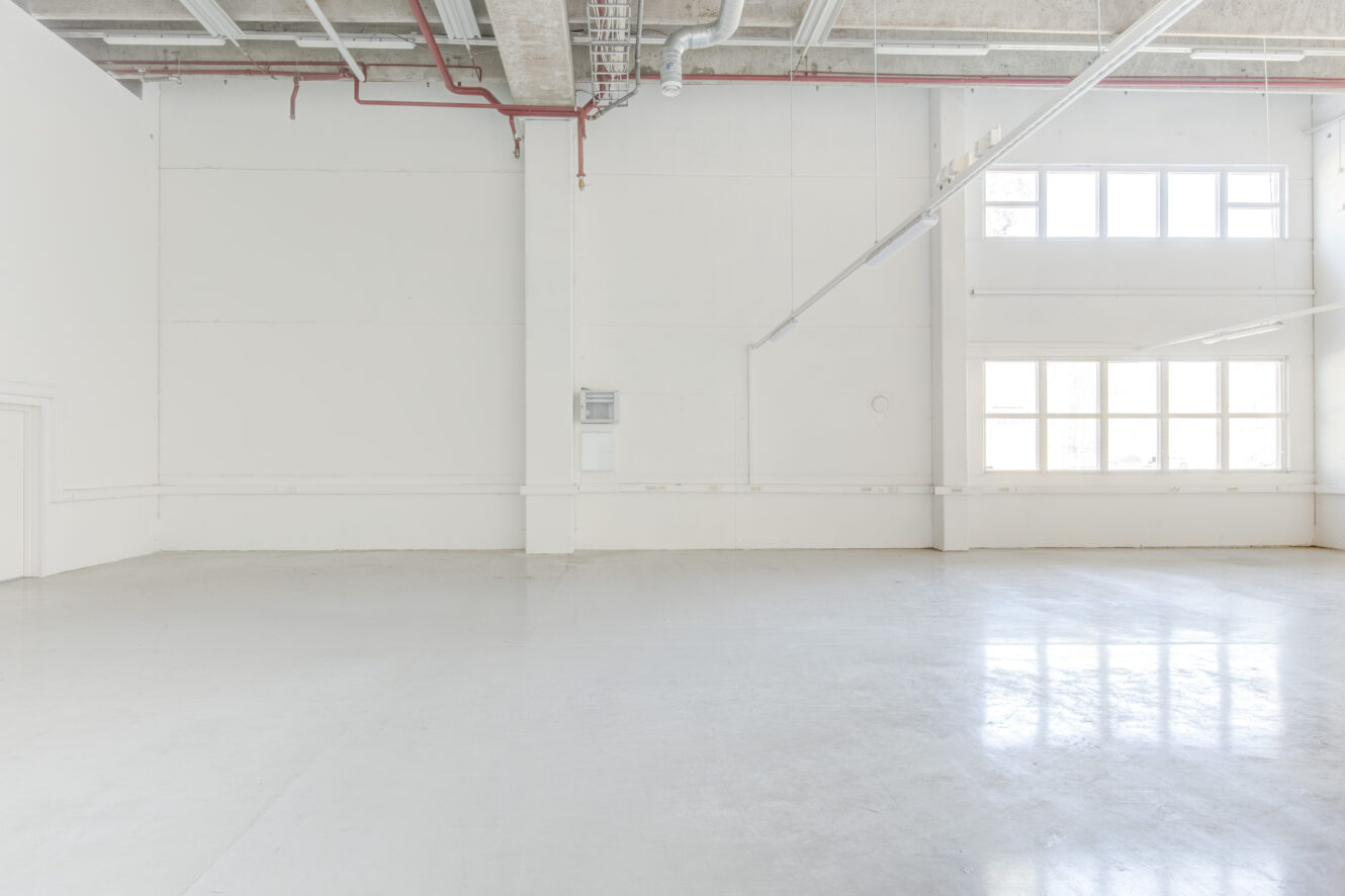 Tilava, tyhjä teollisuushuone, jossa on valkoiset seinät, suuret ikkunat, jotka päästävät luonnonvaloa sisään, ja paljaat kattoputket.