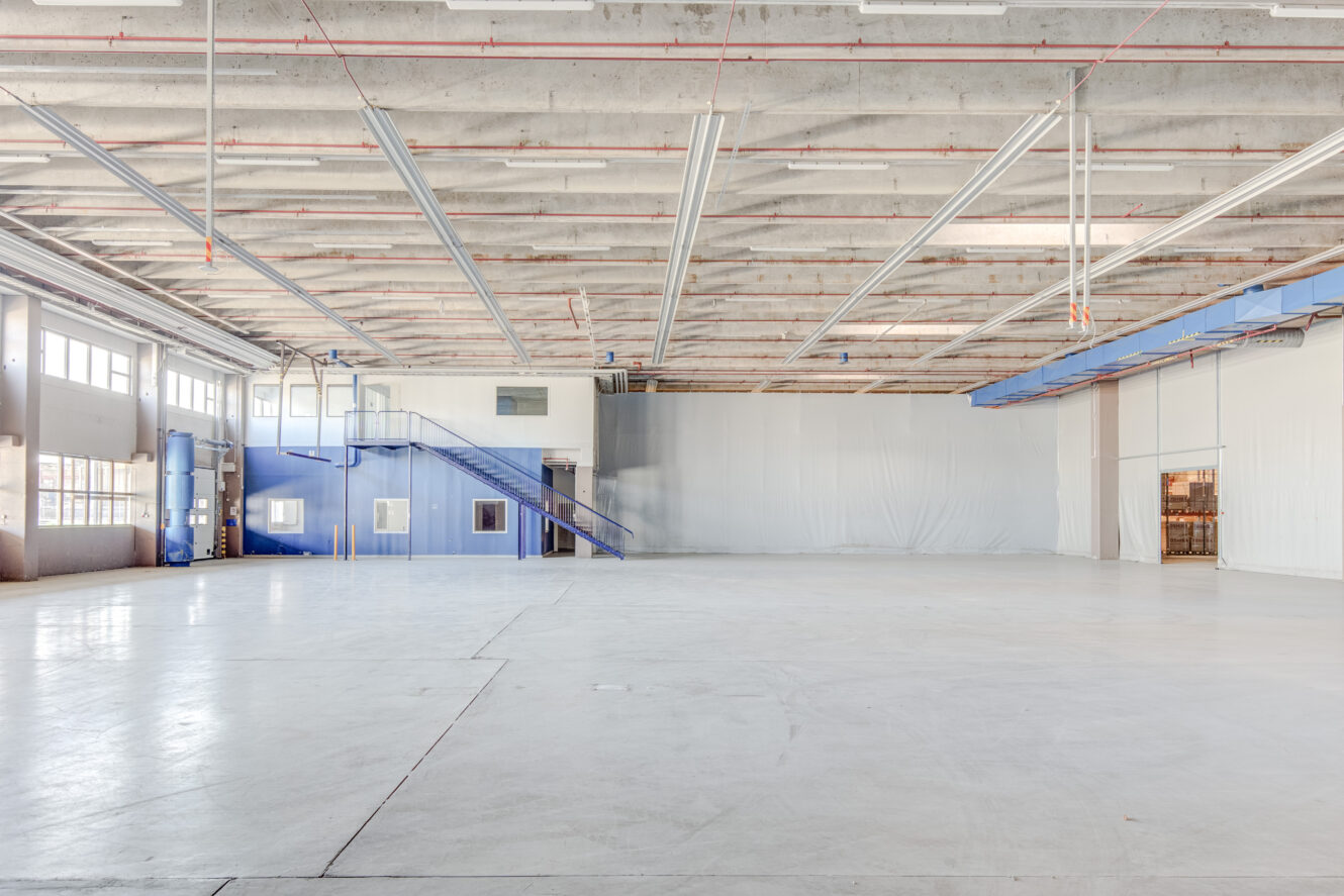 Tilava, tyhjä varaston sisätila, jossa on korkea katto, palkit, suuret ikkunat ja pieni sininen toimistotila, jossa on portaat vasemmalla puolella.