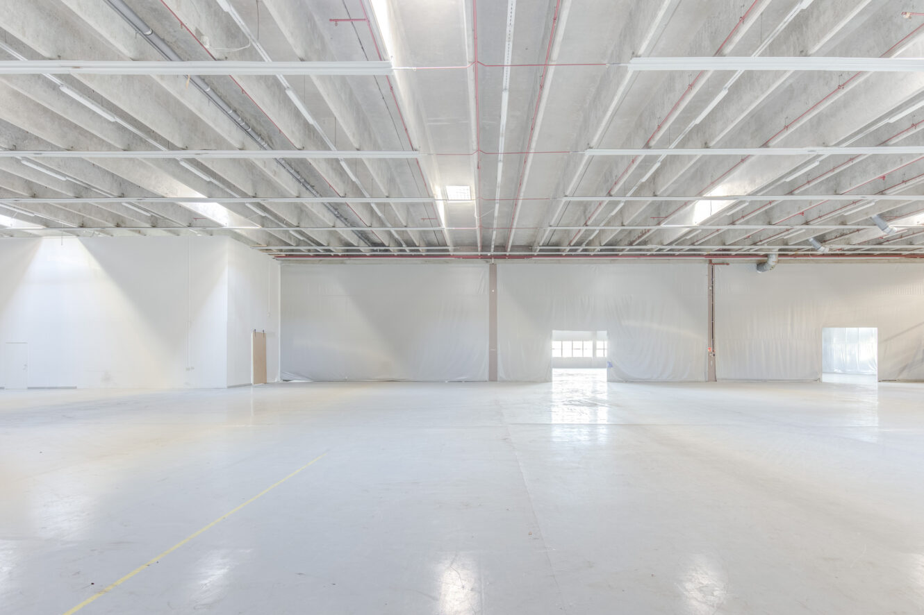 Suuri, tyhjä teollisuusvaraston sisätila, jossa on korkeat katot, palkit, valkoiset seinät ja kirkas valaistus.