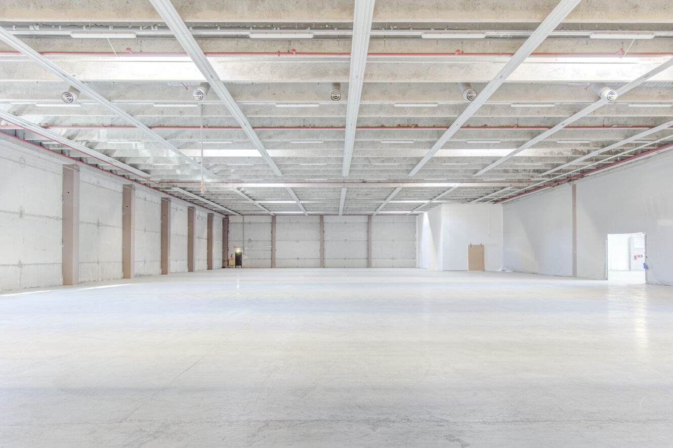 Tyhjä, tilava varastorakennus, jossa on valkoiset seinät, korkea katto ja betonilattia, jota valaisee avoinna olevista oviaukoista tuleva luonnollinen ja keinotekoinen valo.