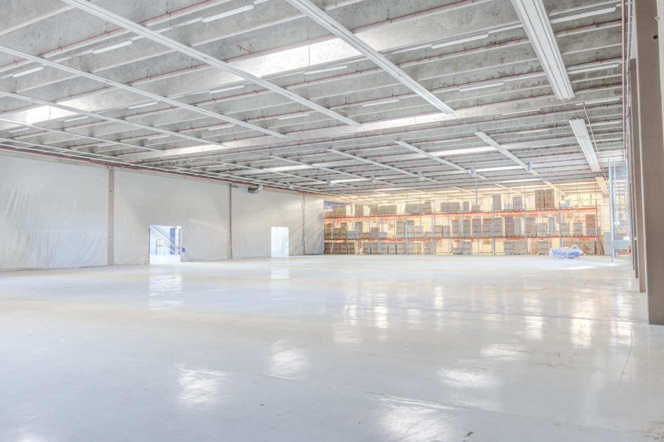 Suuri, tyhjä varastorakennus, jossa on korkeat katot, palkit ja kiiltävä valkoinen lattia; takaseinällä näkyy hyllyjä.