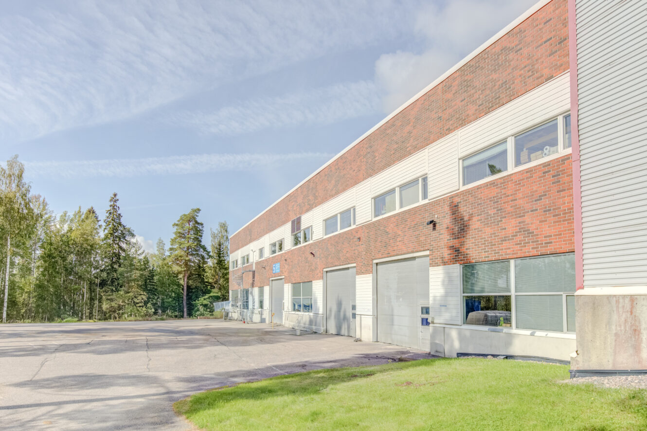 Kaksikerroksinen teollisuusrakennus, jossa on punatiilinen ja beige sivuraide, useita autotallin ovia ja ikkunoita, ja joka sijaitsee päällystetyn alueen vieressä ja puiden ympäröimänä.