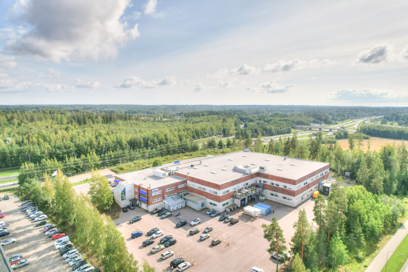 Suuri liikerakennus ja parkkipaikka puiden ja metsän ympäröimänä, taustalla näkyy valtatie ja autoja osittain pilvisen taivaan alla.