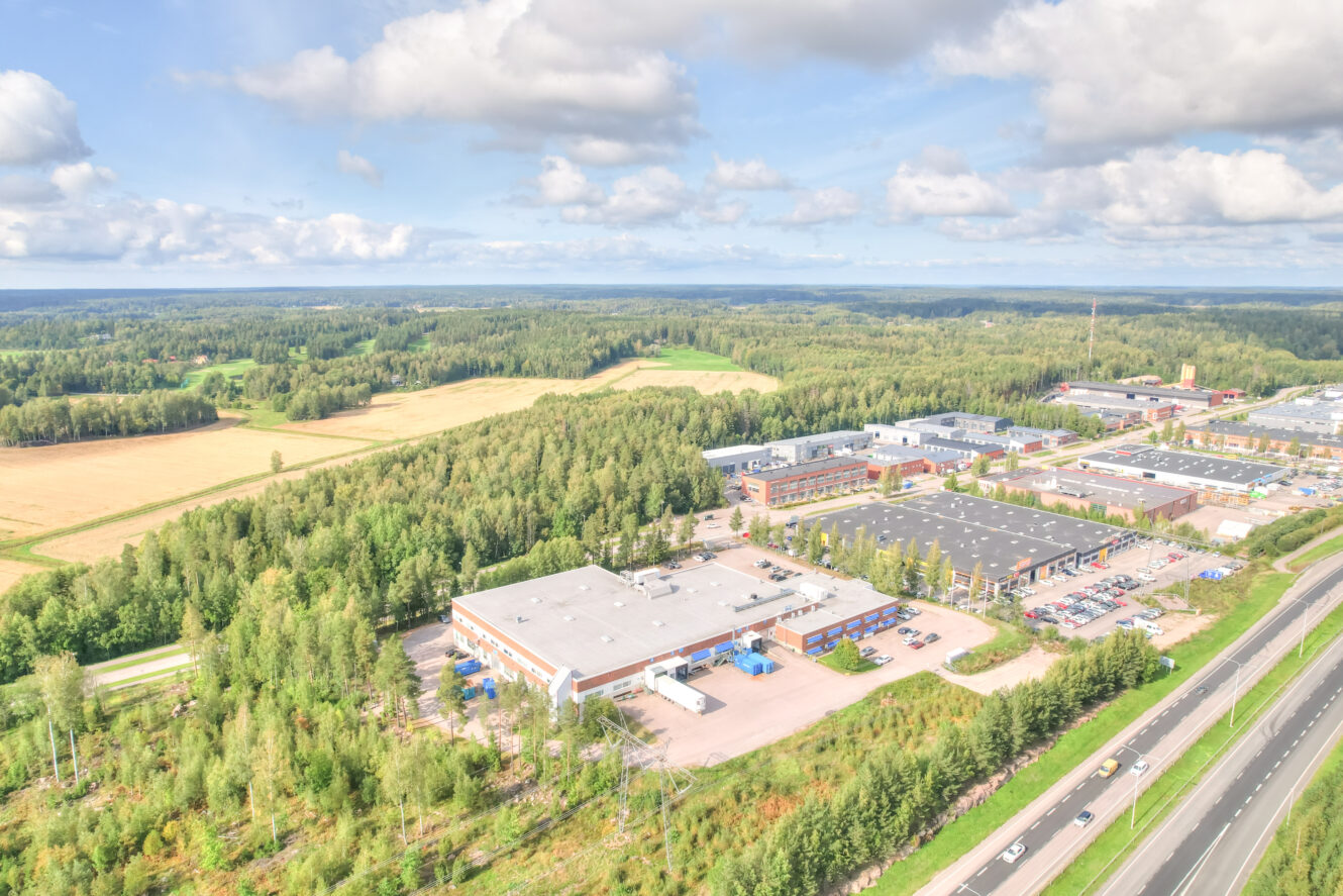 Ilmakuva liikerakennuksista ja pysäköintialueista valtatien vieressä, metsän ja peltojen ympäröimänä osittain pilvisen taivaan alla.
