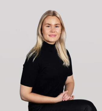 Henkilö, jolla on pitkät vaaleat hiukset ja päällään musta lyhythihainen poolopaita, istuu kädet ristissä kameraan päin vaaleanharmaata taustaa vasten.