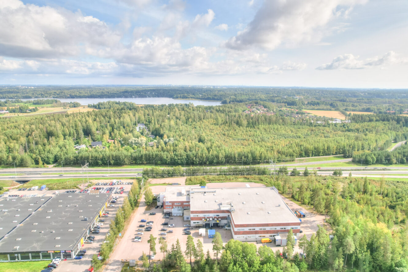 Ilmakuva teollisuusalueesta, jossa on varastoja, parkkipaikkoja ja autoja, metsän ja peltojen ympäröimänä, taustalla järvi ja kaukaisia rakennuksia osittain pilvisen taivaan alla.