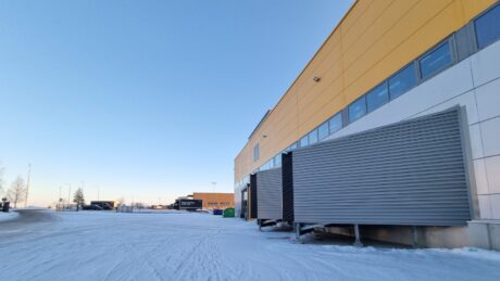 Suuri teollisuusrakennus, jonka seinät ovat keltaiset ja valkoiset, lastauslaiturin ovet ovat kiinni ja lumi peittää maan kirkkaalla taivaalla.