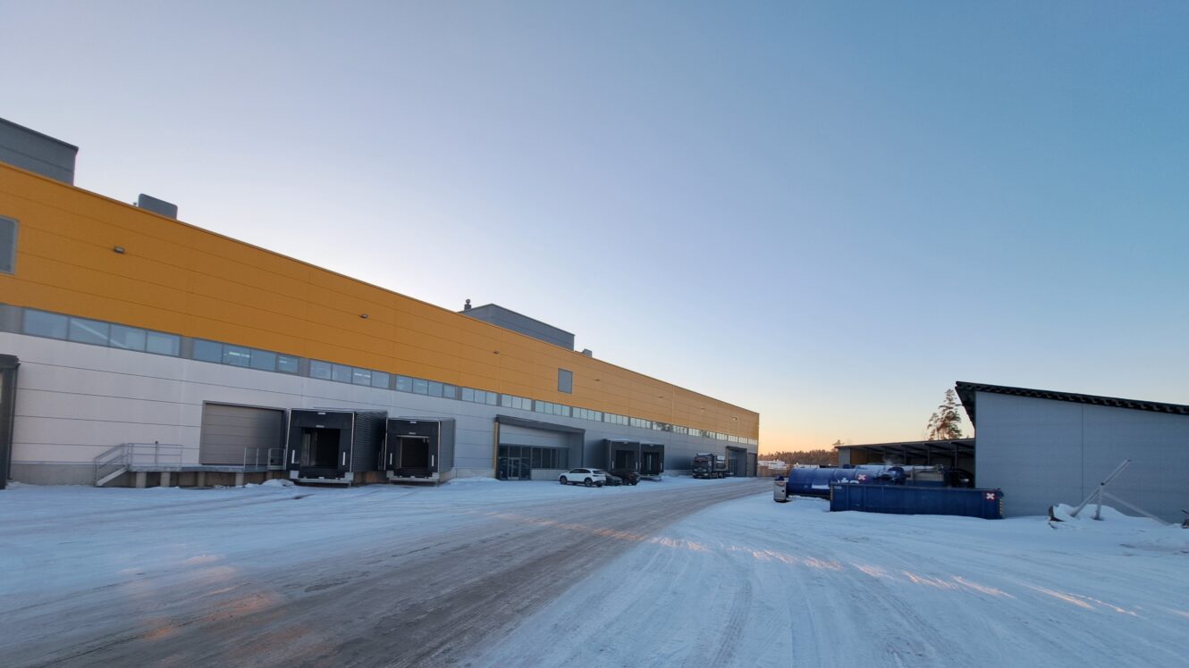 Suuri teollisuusvarasto, jonka julkisivu on keltainen ja harmaa, useita lastauslaitureita, pysäköityjä kuorma-autoja ja lunta, joka peittää maan kirkkaalla taivaalla.