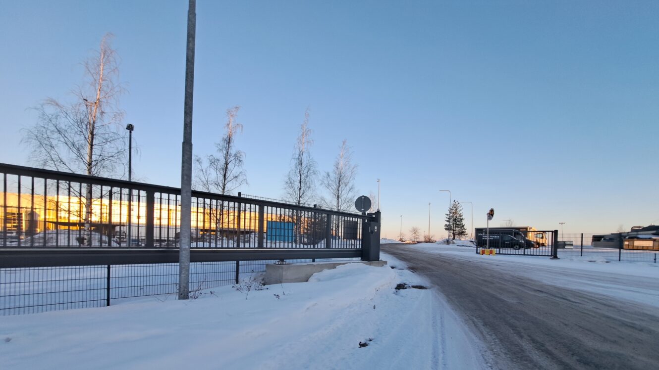 Lumipeitteinen tie kulkee korkean metalliaidan vieressä, ja taustalla näkyy paljaita puita ja pysäköityjä ajoneuvoja kirkkaalla taivaalla auringonlaskun aikaan.