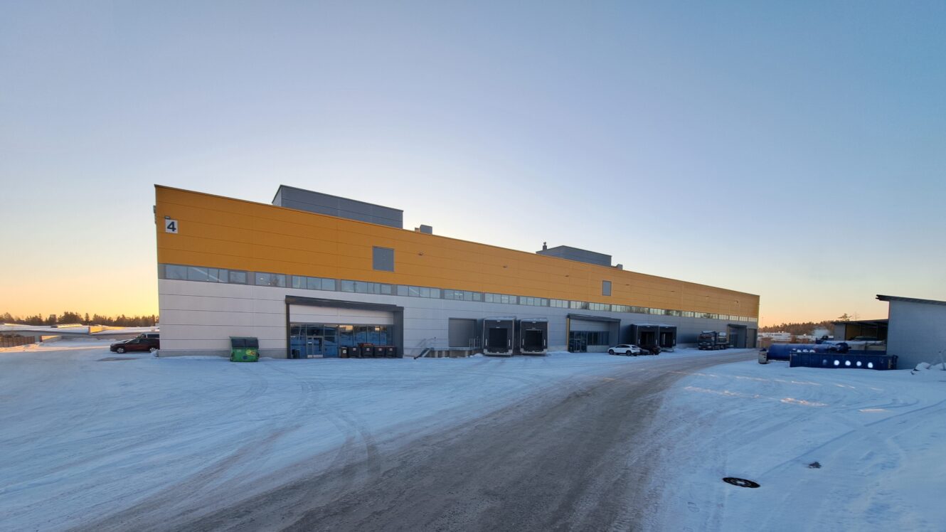 Suuri teollisuusvarasto, jossa on keltainen yläjulkisivu ja useita lastauslaiturin ovia, lumen ympäröimänä kirkkaalla taivaalla auringonnousun tai -laskun aikaan.