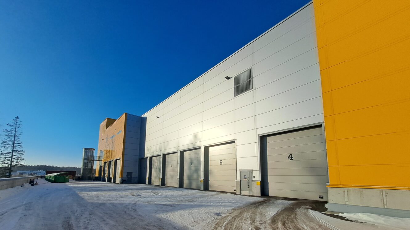 Rivi suuria teollisuuslaitoksen lastauslaiturin ovia varastorakennuksessa talvella, kun maassa on lunta ja taivas on kirkkaansininen.