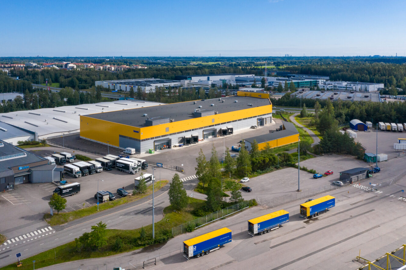 Suuri keltaisella katolla varustettu varasto, jossa on useita lastauslaitureita, kuorma-autoja ja perävaunuja, sijaitsee teollisuusalueella puiden ja muiden rakennusten ympäröimänä.