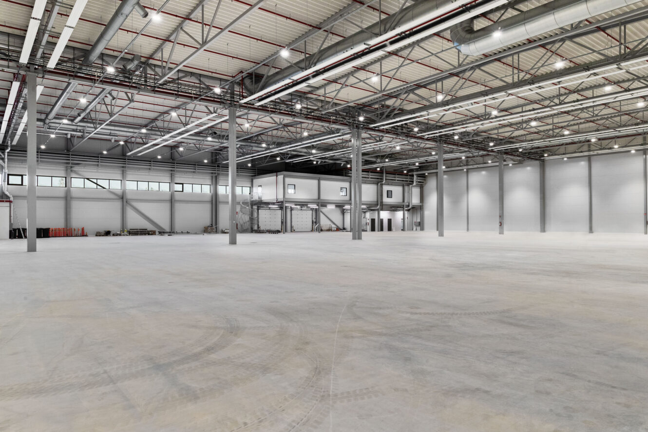Tilava, tyhjä varaston sisätila, jossa on korkeat katot, paljaat rakennepalkit ja useita ikkunoita yläseinillä.