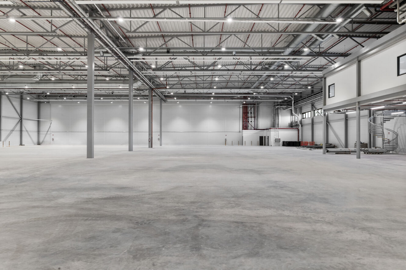 Tilava, tyhjä teollisuusvarasto, jossa on betonilattia, korkea katto, palkit ja minimaalinen sisustus.