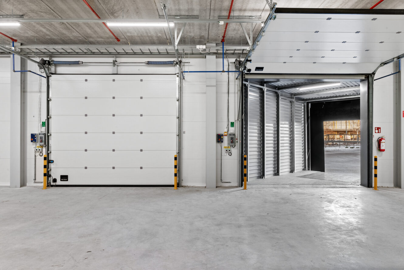 Puhdas, tyhjä teollisuusvarasto, jossa on kaksi suurta lastauslaiturin ovea, joista toinen on suljettu ja toinen auki, ja sen takaa avautuu ulkoalue.