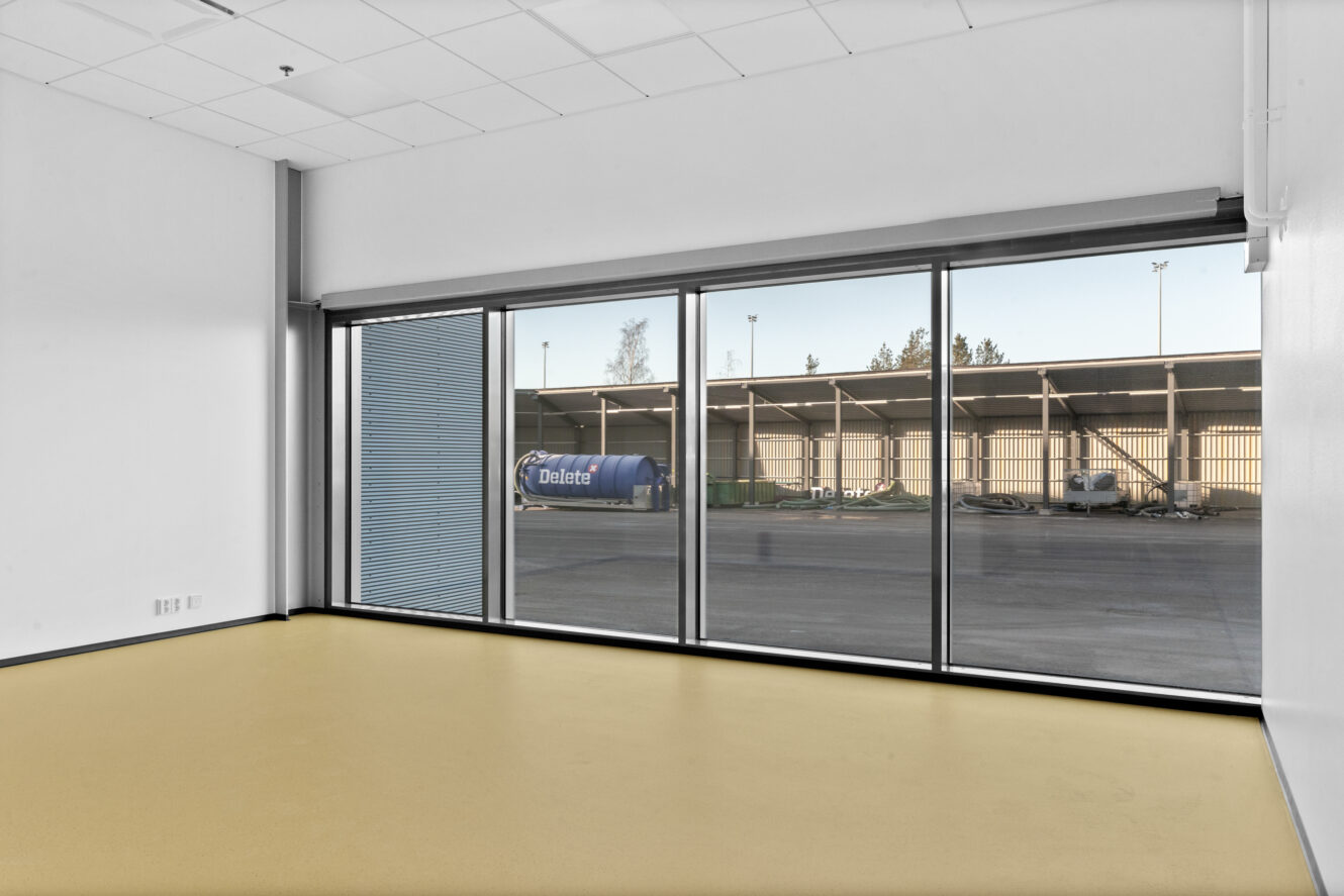 Tyhjä huone, jossa on keltainen lattia ja suuret lattiasta kattoon ulottuvat ikkunat, joista avautuu näkymä teollisuusalueelle, jossa on kuorma-auto, jossa lukee Delete.