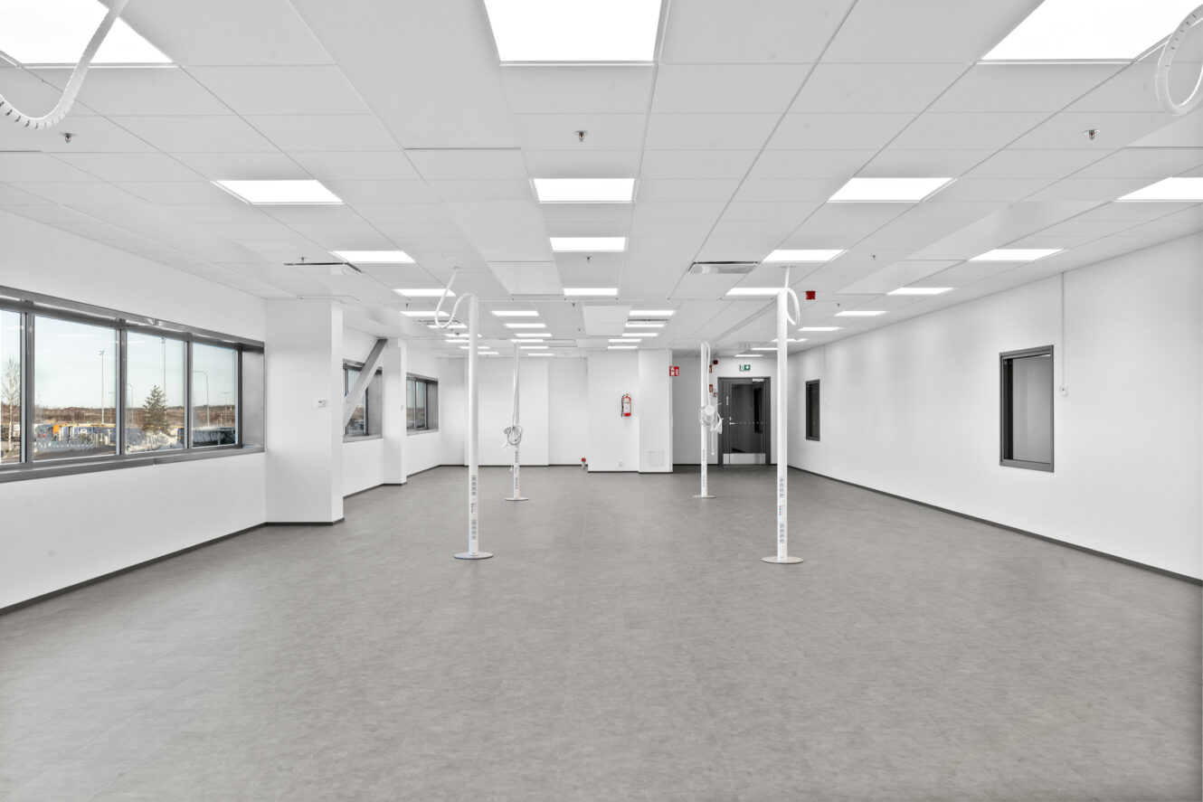 Suuri, tyhjä, hyvin valaistu huone, jossa on valkoiset seinät, suuret ikkunat vasemmalla, harmaa lattia ja useita kattoon kiinnitettyjä valaisimia.