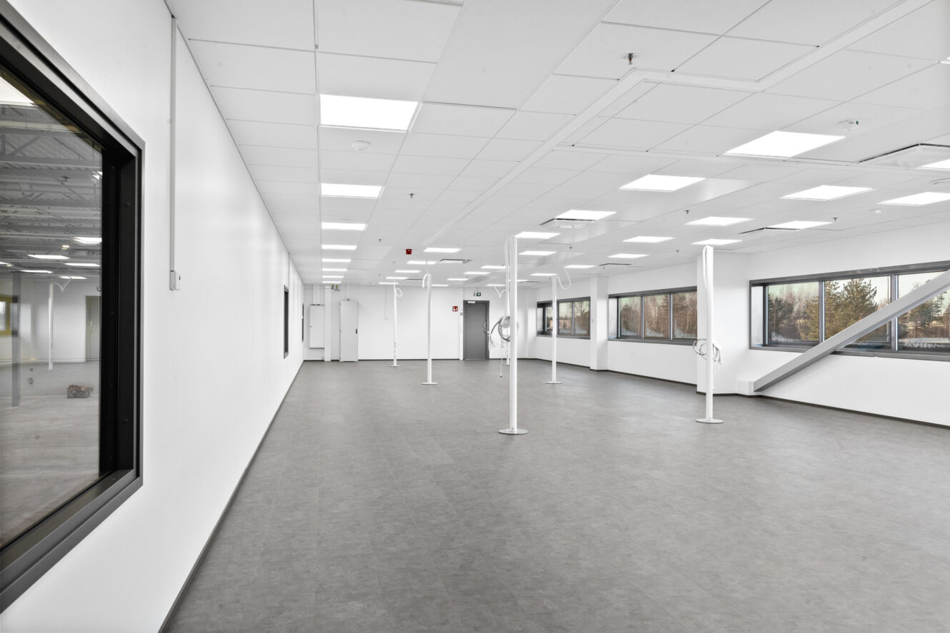 Suuri, tyhjä huone, jossa on valkoiset seinät, harmaa lattia, kattovalaisimet, tukipylväät ja useita suuria ikkunoita sivuilla.