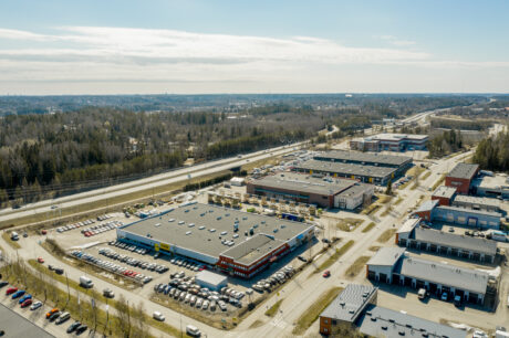 Ilmakuva kaupallisesta alueesta, jossa on useita suuria rakennuksia, autoja täynnä olevia parkkipaikkoja ja lähellä kulkeva valtatie, jota ympäröivät puut ja tiet.