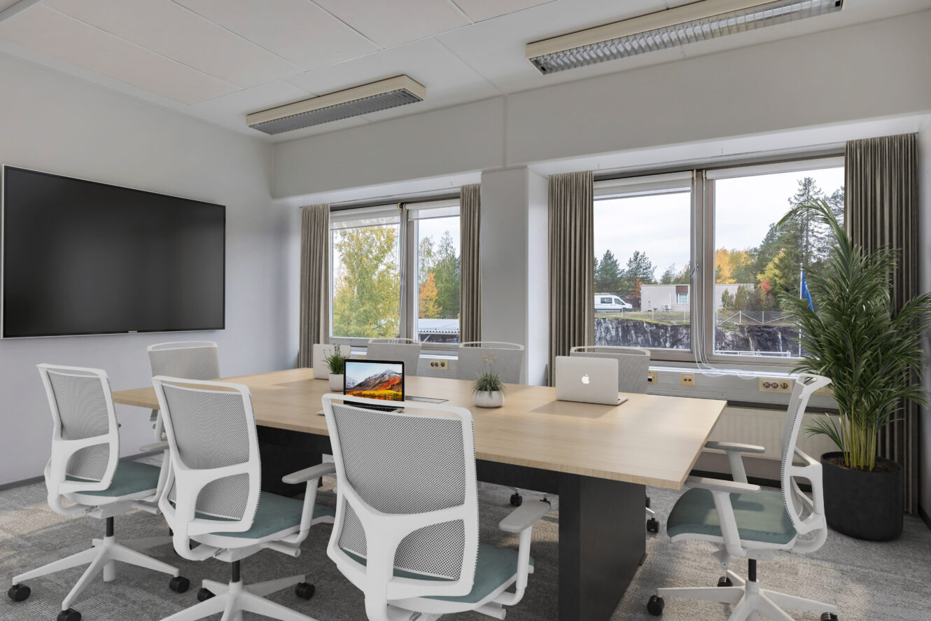 Moderni kokoushuone, jossa on puinen pöytä, kuusi valkoista toimistotuolia, suuri seinälle kiinnitetty näyttö, kannettavia tietokoneita, ruukkukasveja ja suuret ikkunat, joista näkyy puita ulkona.