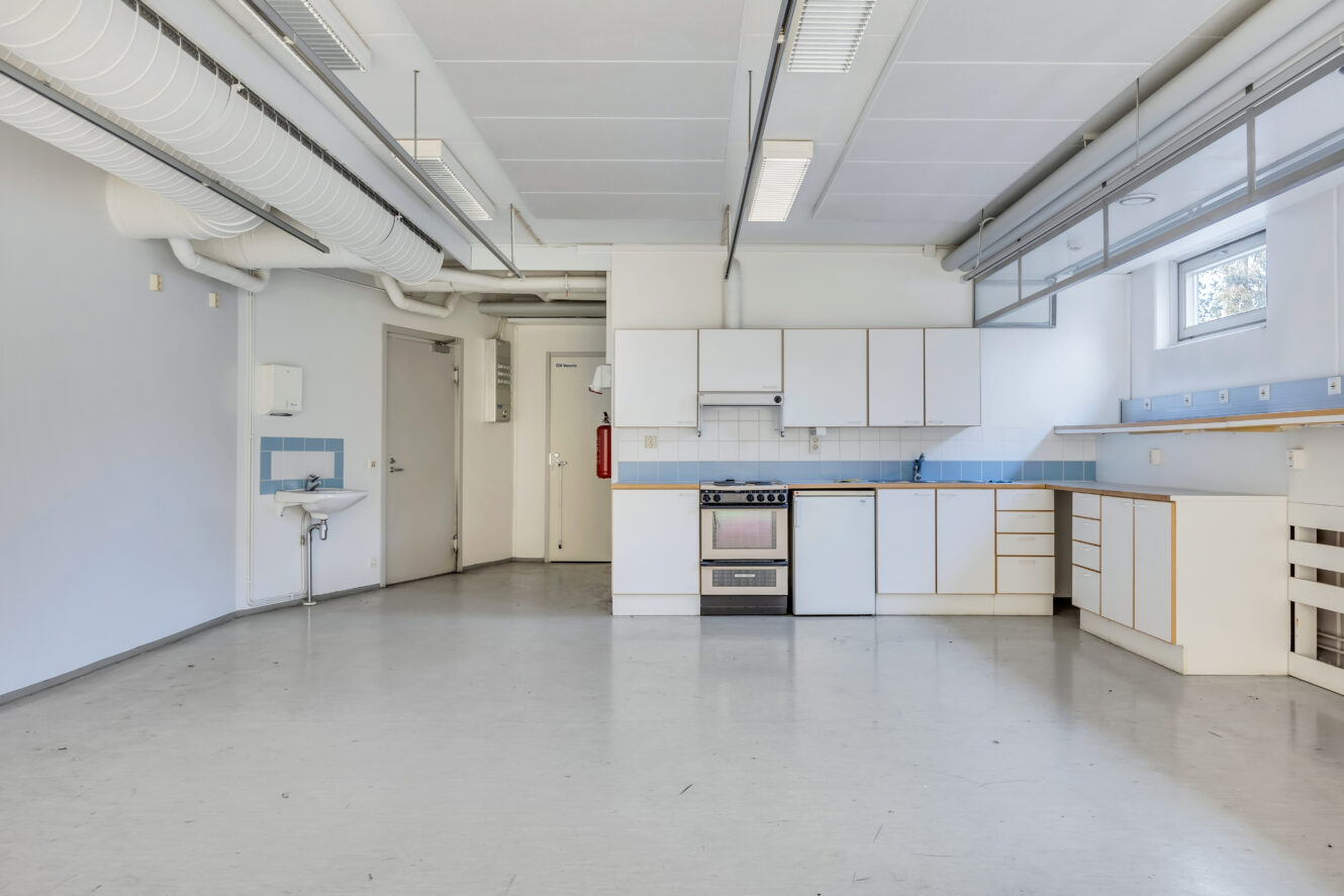 Tilava, tyhjä keittiö, jossa on valkoiset kaapit, liesi, allas ja laatoitettu välitilalevy huoneessa, jossa on vaaleanharmaat lattiat ja teollisuustyyliset kattovalaisimet.