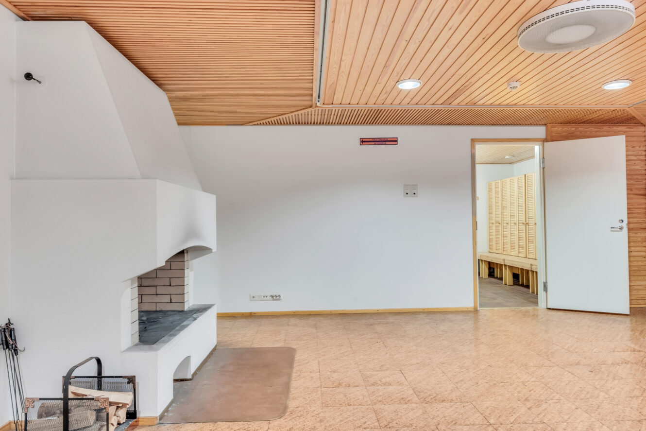 Huone, jossa on valkoinen takka, puinen katto, korkkilattia ja avoin ovi, joka johtaa tilaan, jossa on puisia kaappeja.