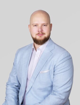 Vaaleansiniseen puvun takkiin ja vaaleaan paitaan pukeutunut kalju, parrakas mies istuu vaaleanharmaata taustaa vasten.