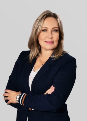 Vaaleatukkainen nainen, jolla on tummansininen bleiseri ja valkoinen toppi, seisoo kädet ristissä vaaleanharmaata taustaa vasten.