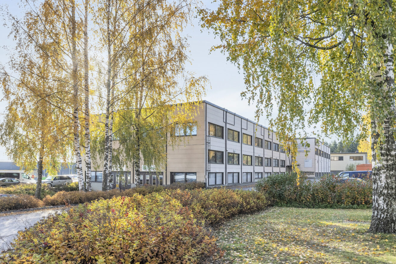 Moderni kaksikerroksinen toimistorakennus, jossa on suuret ikkunat, on syksyisten puiden ja pensaiden ympäröimä, ja taustalla näkyy pysäköityjä autoja.