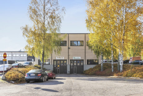 Beige kaksikerroksinen liikerakennus, jonka edessä on kylttejä ja useita pysäköityjä autoja, ja jota ympäröivät keltaisia syyslehtiä kasvavat koivut.