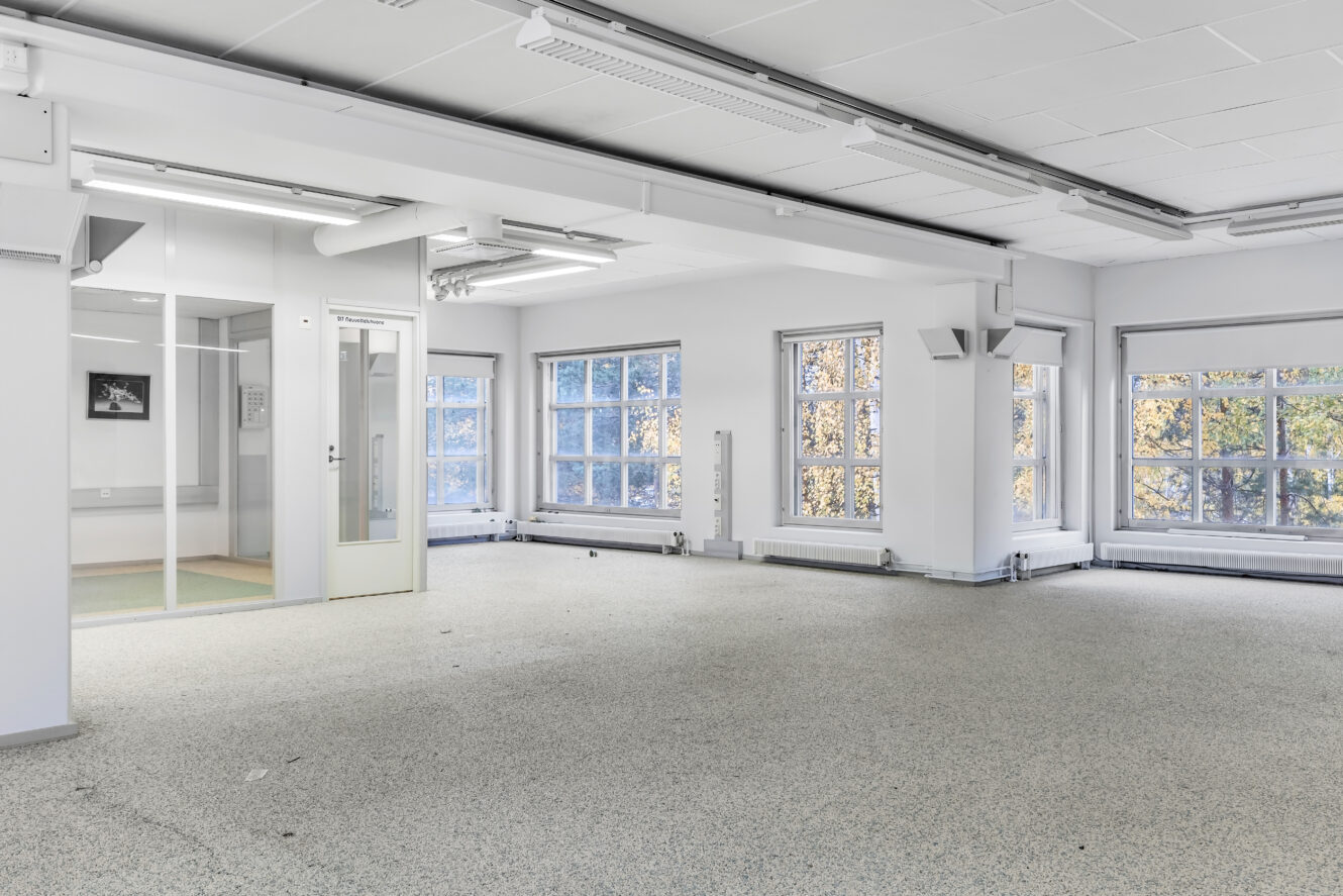 Tilava, tyhjä toimistohuone, jossa on suuret ikkunat, valkoiset seinät ja harmaa laikullinen lattia. Vasemmalla puolella näkyy lasiseinäinen huone ja ovi. Luonnonvalo täyttää tilan.