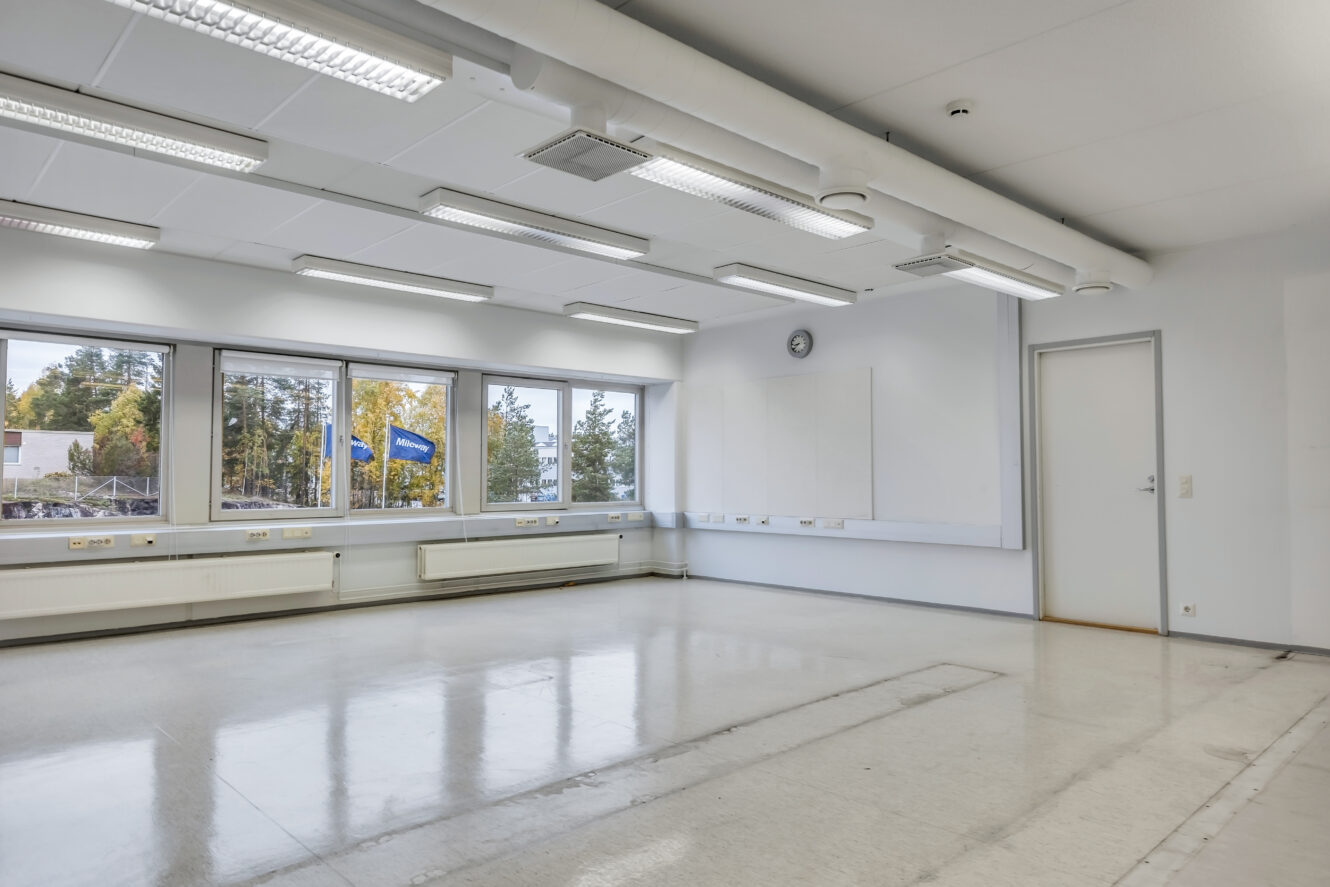 Tyhjä luokkahuone, jossa on suuret ikkunat, loisteputkivalot, valkoiset seinät, kello ja suljettu ovi. Lattian heijastukset osoittavat, että huone on puhdas ja hyvin valaistu.