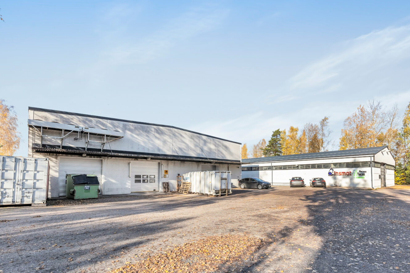 Teollisuusrakennuksia, sorapintainen pysäköintialue, useita autoja, kuljetuskontteja ja hajallaan olevia kuormalavoja, syksyisten puiden ympäröimänä.