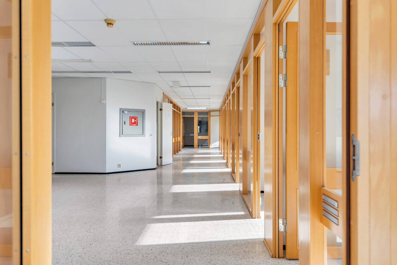 Tyhjä käytävä modernissa toimistorakennuksessa, jossa on lasiseinäisiä huoneita ja puukehyksiä, ja jota valaisee ikkunoista tuleva luonnonvalo.