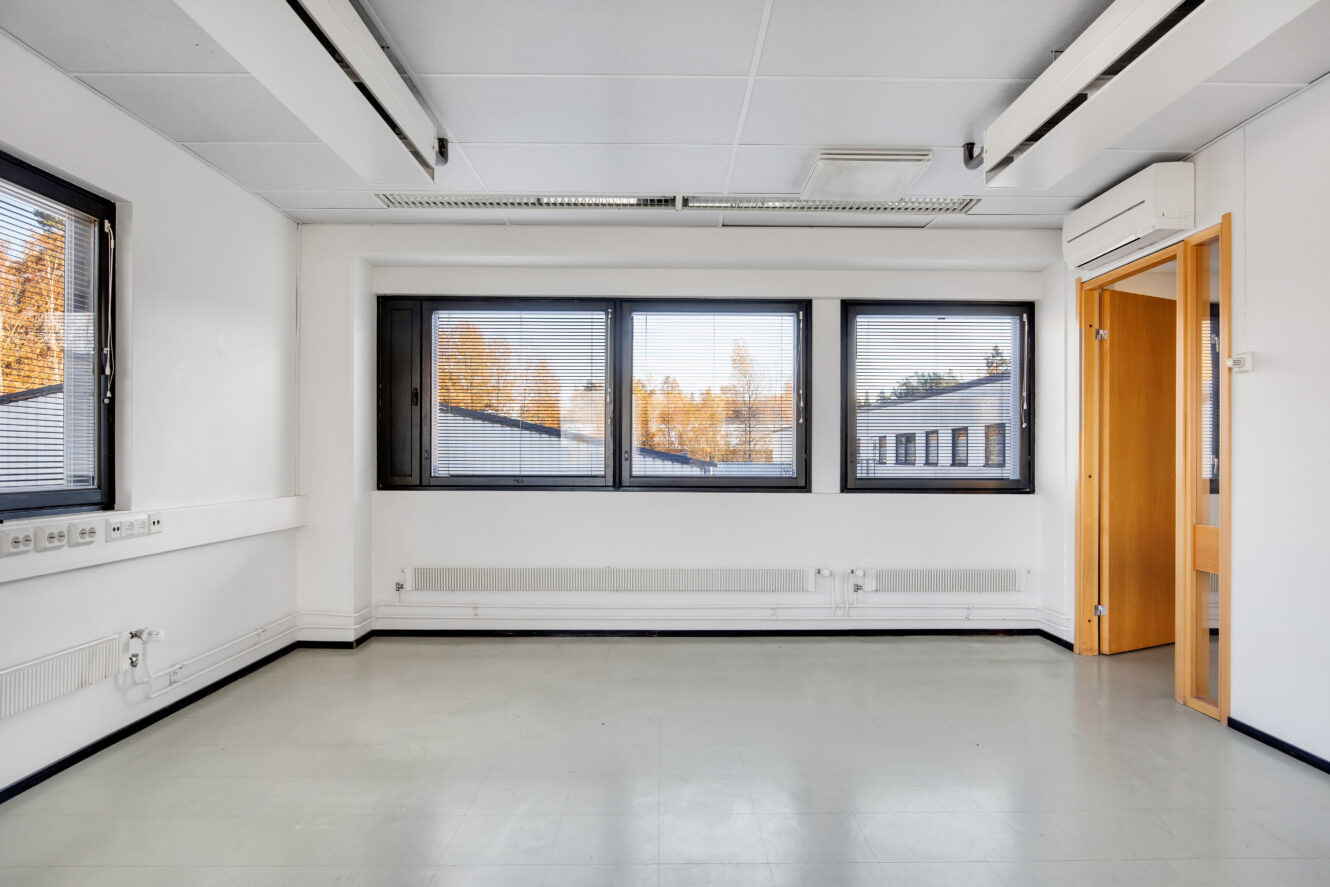 Tyhjä toimistohuone, jossa on valkoiset seinät, kolme suurta ikkunaa, joissa on kaihtimet, ylävalaistus, vaaleanharmaa lattia ja avoin puuovi.
