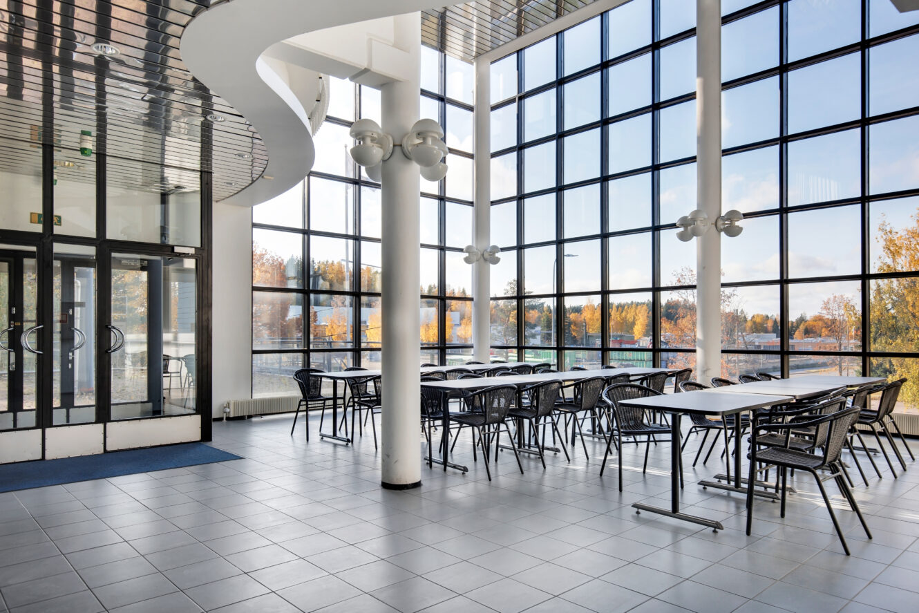 Moderni kahvila, jossa on suuret lattiasta kattoon ulottuvat ikkunat, mustat tuolit ja valkoiset pöydät, luonnonvalo valaisee ja josta on näkymät syyslehtisten puiden yli.