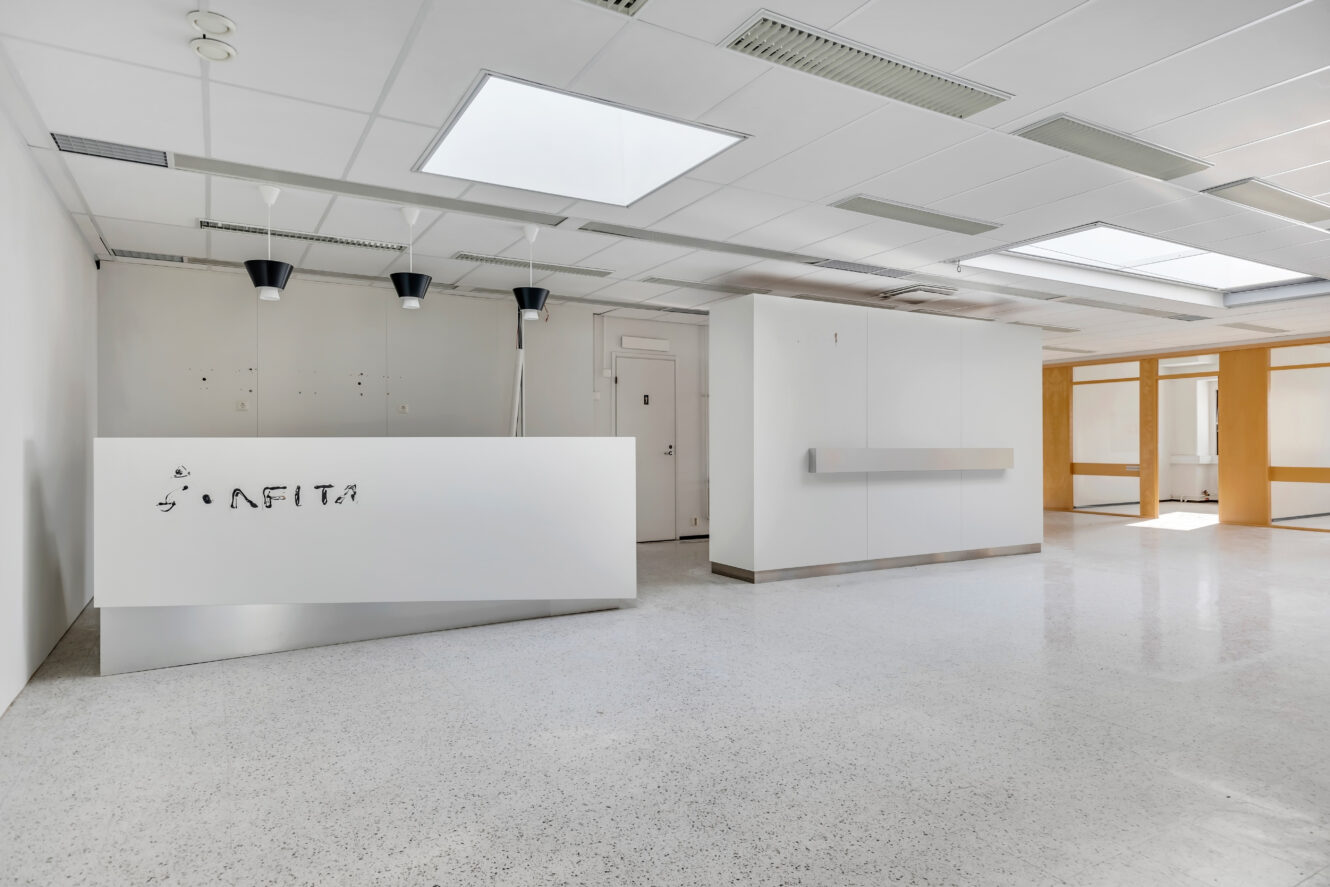 Tyhjä moderni vastaanottotila, jossa on valkoinen työpöytä, riippuvalaisimet, valkoiset seinät ja suuret ikkunat, jotka päästävät luonnonvaloa tilaan.