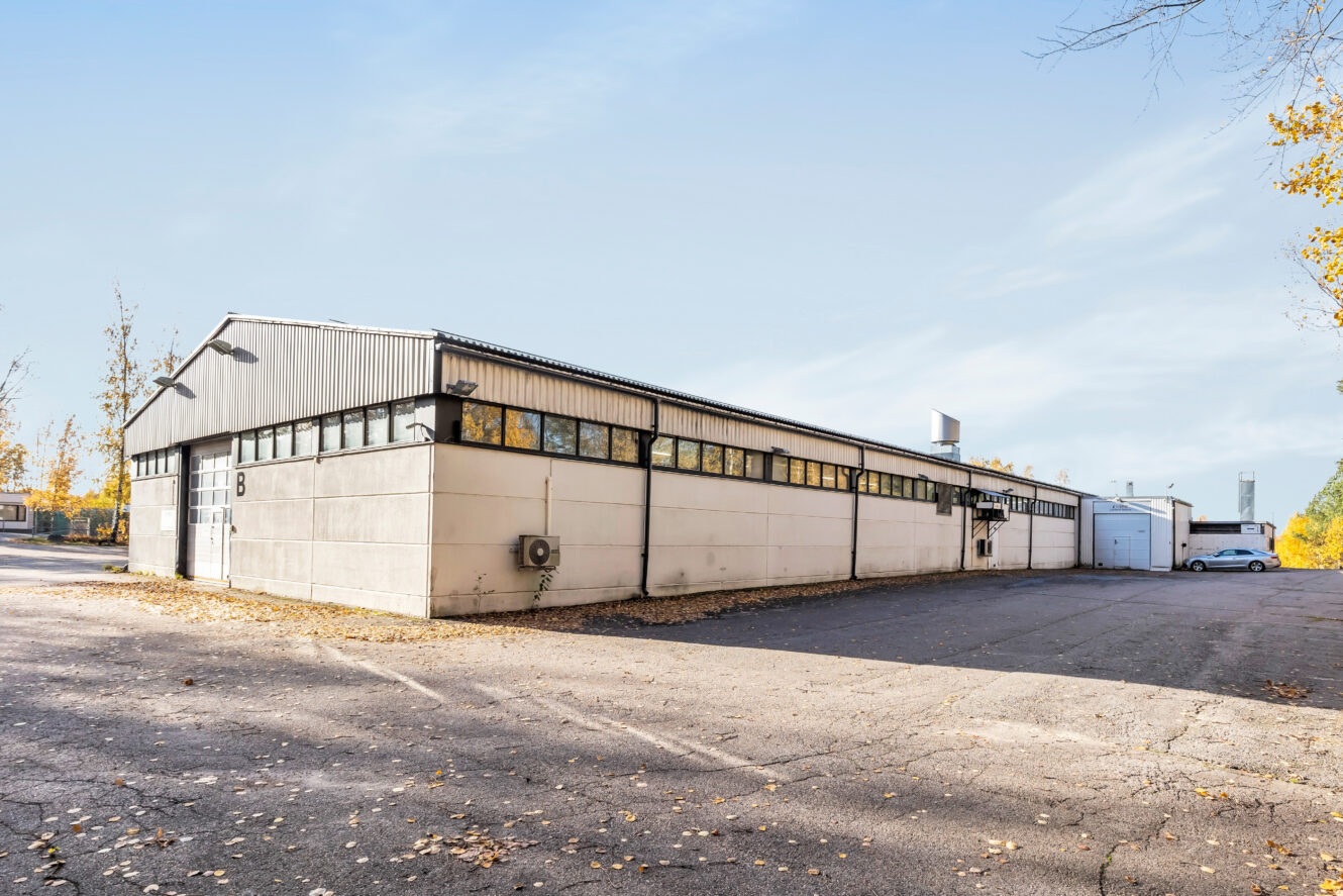 Yksikerroksinen teollisuusvarasto, jossa on metallikatto, valkoiset ulkoseinät ja useita ikkunoita ja joka sijaitsee asfalttialueella, jossa on hajallaan syksyn lehtiä.