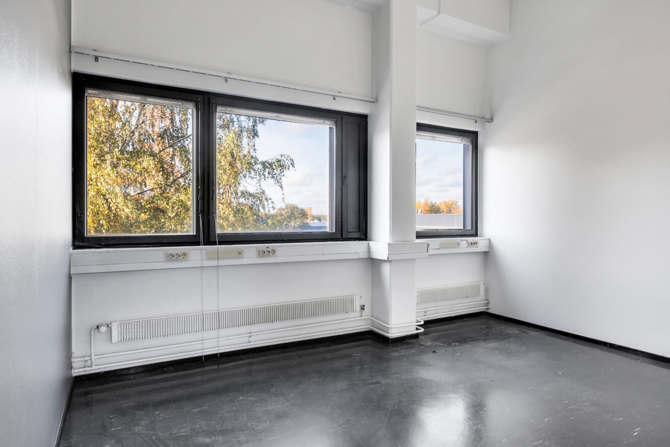 Tyhjä huone, jossa on musta lattia, valkoiset seinät, suuret ikkunat, joista näkyy puita ja taivas, ja patterit ikkunoiden alla.