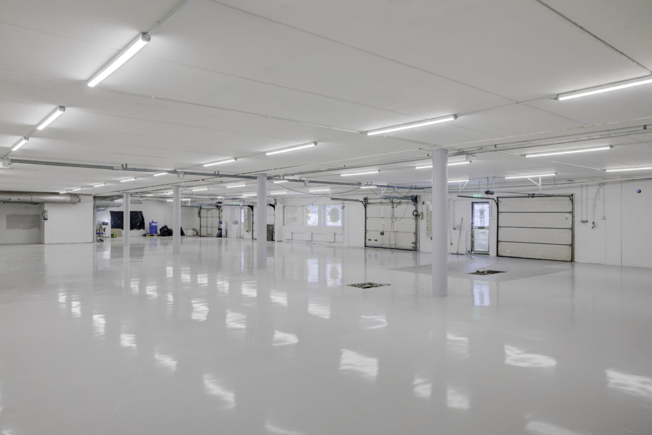 Suuri, tyhjä teollisuustila, jossa on valkoiset seinät, kiiltävät lattiat, loisteputkivalot ja useita autotallin ovia.