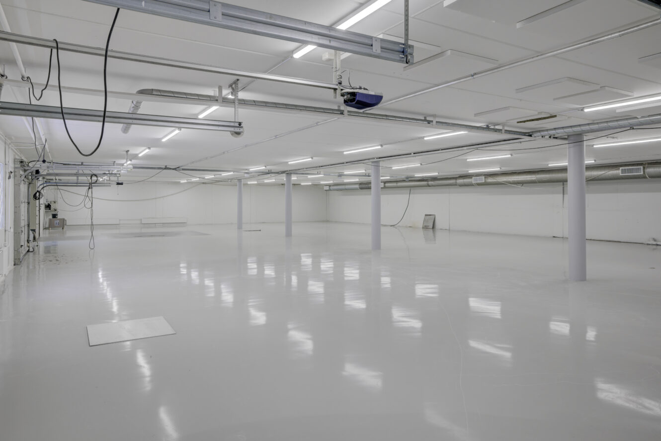 Suuri, tyhjä teollisuusvarastotila, jossa on valkoiset lattiat, valkoiset seinät, kattovalaisimet, tukipylväät sekä paljaat putket ja johdot.
