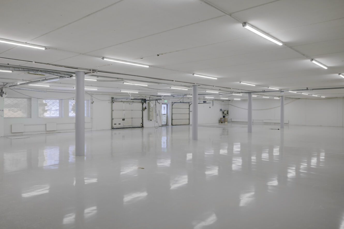 Suuri tyhjä teollisuusvarasto, jossa on valkoiset seinät, kiillotettu lattia, kattovalaisimet, paljaat putket ja kaksi autotallin ovea.