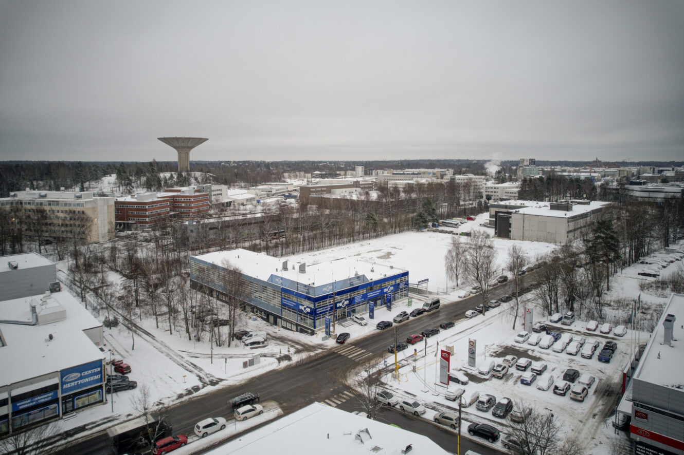 Luminen kaupunkialue, jossa on liikerakennuksia, pysäköityjä autoja ja taustalla näkyy suuri vesitorni pilvisellä taivaalla.